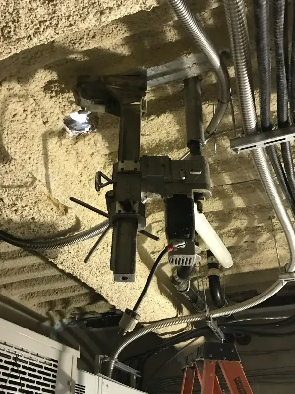 A heavy-duty core drill mounted on a ceiling stand, surrounded by exposed pipes and textured spray-on fireproofing.