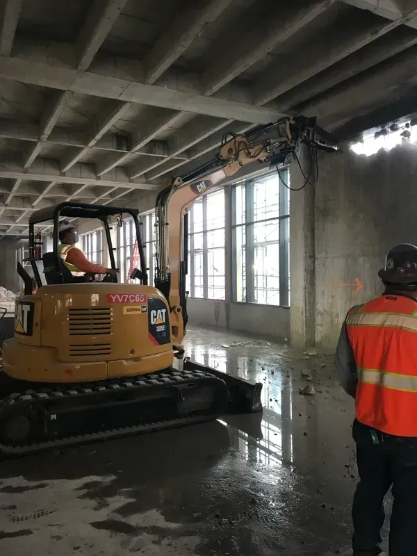A construction worker operates a yellow CAT excavator to break a concrete wall inside an unfinished, water-flooded building.