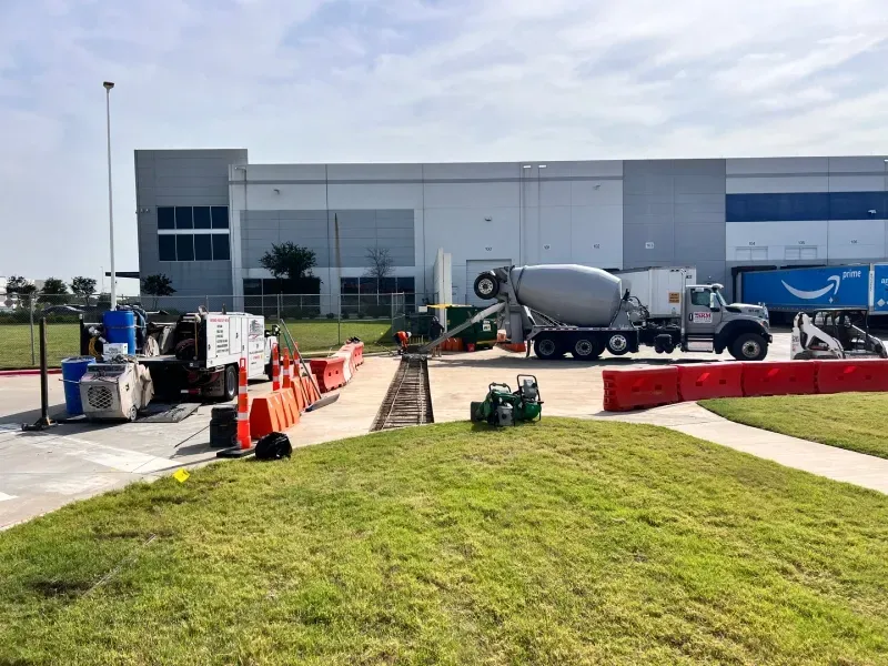 A concrete mixer truck pours concrete into a trench at a construction site in front of a large Amazon warehouse.