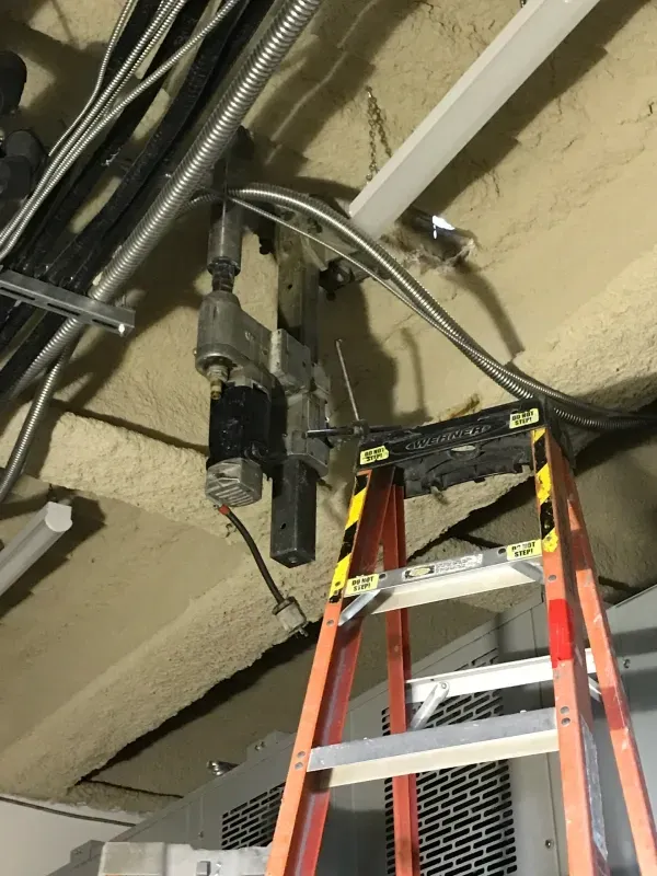 A mechanical actuator hangs from a ceiling near a ladder in a utility space with exposed wires and foam insulation.
