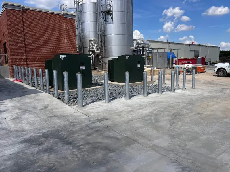 Two green utility cabinets behind a line of bollards on a concrete pad near a brick building and large metal silos.