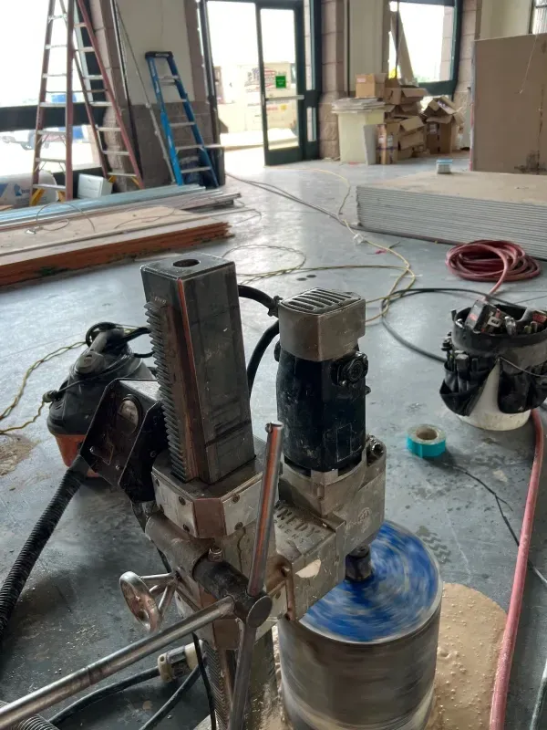 A industrial core drilling machine cuts into a concrete floor at a construction site with ladders in the background.