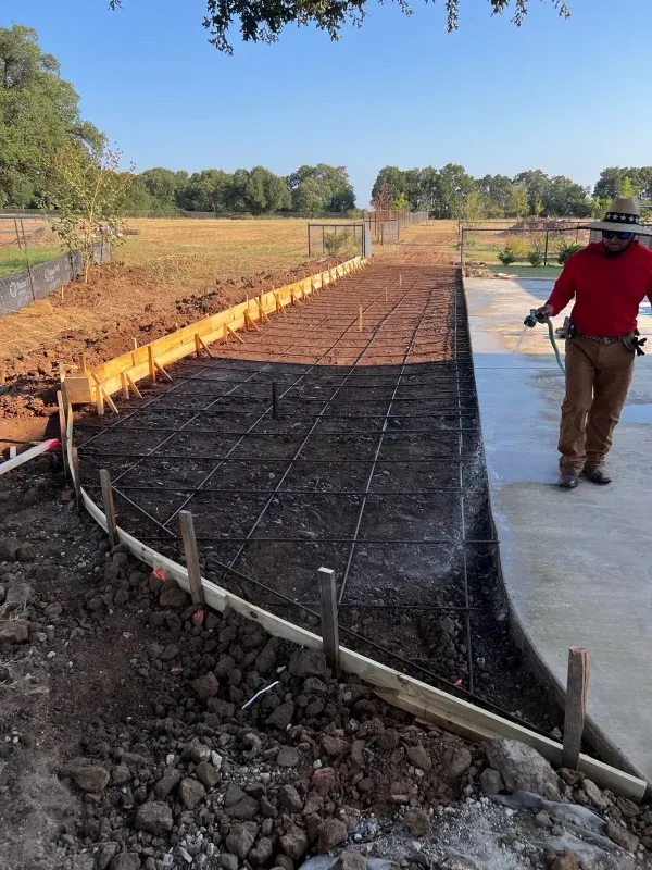 A person in a red shirt and hat stands near a wooden-formed construction site with a wire mesh grid set for concrete.