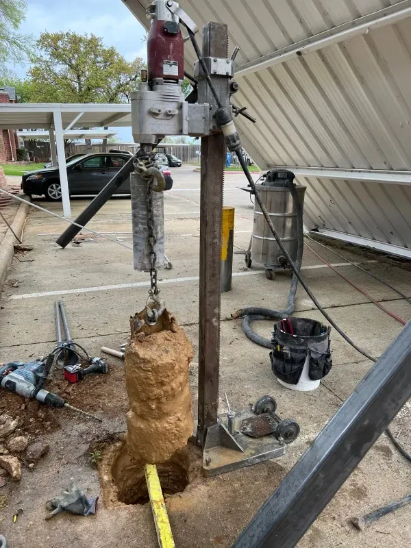 A heavy-duty drill stand mounted on concrete, extracting a large, cylindrical core sample from the ground outside.