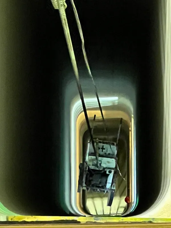 A small, white, vintage-style vehicle is suspended by cables inside a narrow, vertical light well or stairwell shaft.