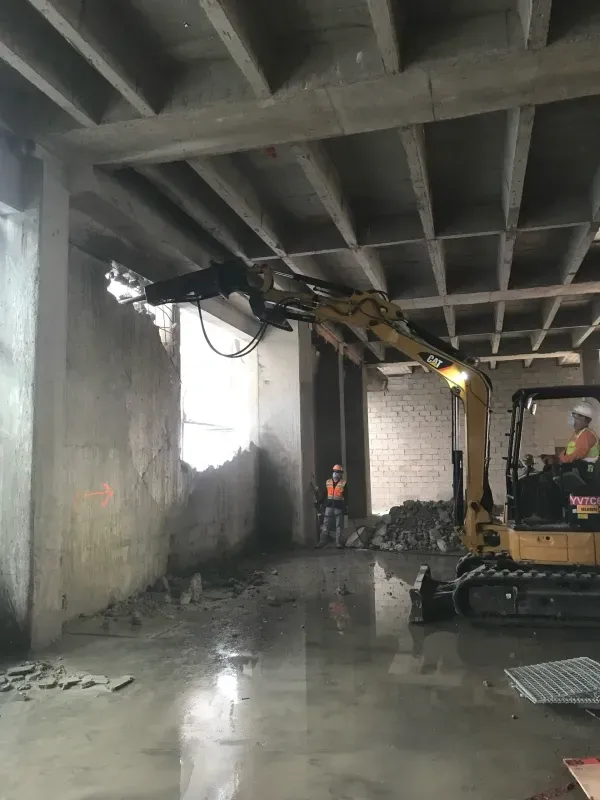 A yellow excavator with a hydraulic breaker attachment demoing a concrete wall inside an unfinished building.