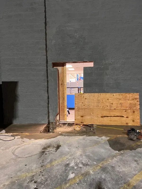 An unfinished doorway cut into a grey brick exterior wall, partially boarded up with plywood, revealing an interior room.