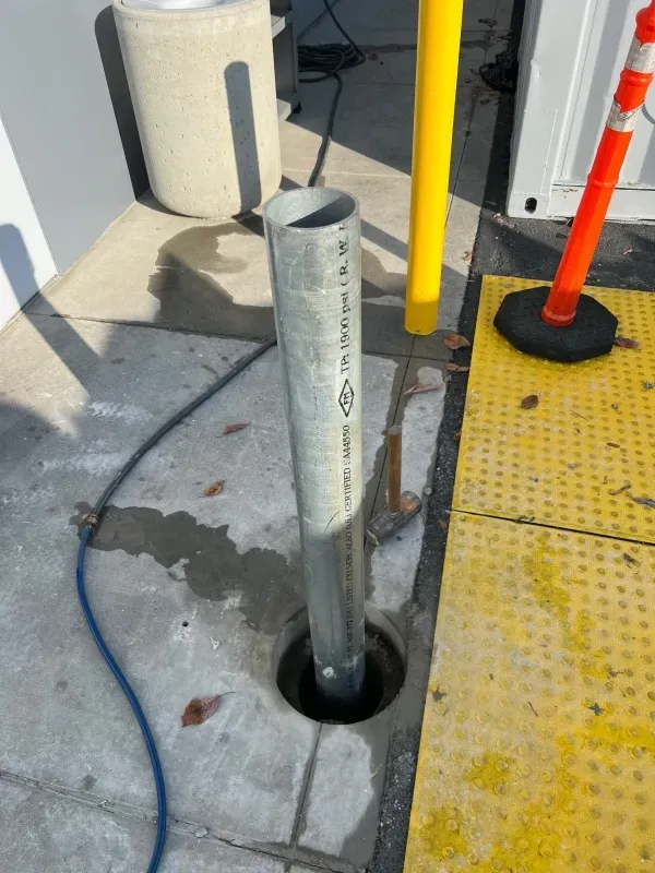 A metal post sits in a concrete hole near a yellow tactile paving strip, next to a yellow bollard and orange traffic cone.