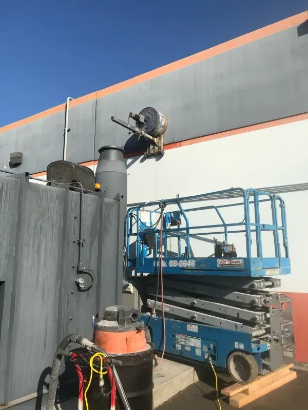 A blue scissor lift stands next to a gray industrial unit with a vent stack and a black canister under a clear blue sky.