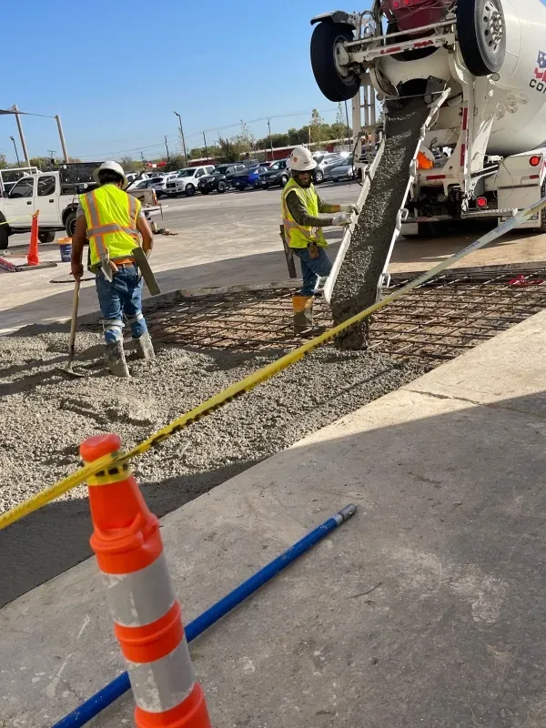 Two workers in high-visibility vests and hard hats pour wet concrete from a mixer truck onto a prepared gravel surface.