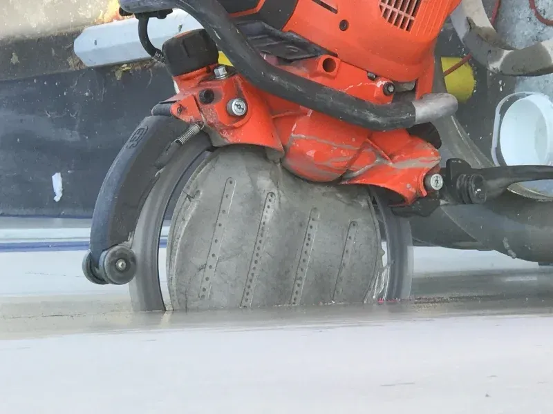 A close-up view of an orange concrete saw cutting into a flat, gray paved surface.