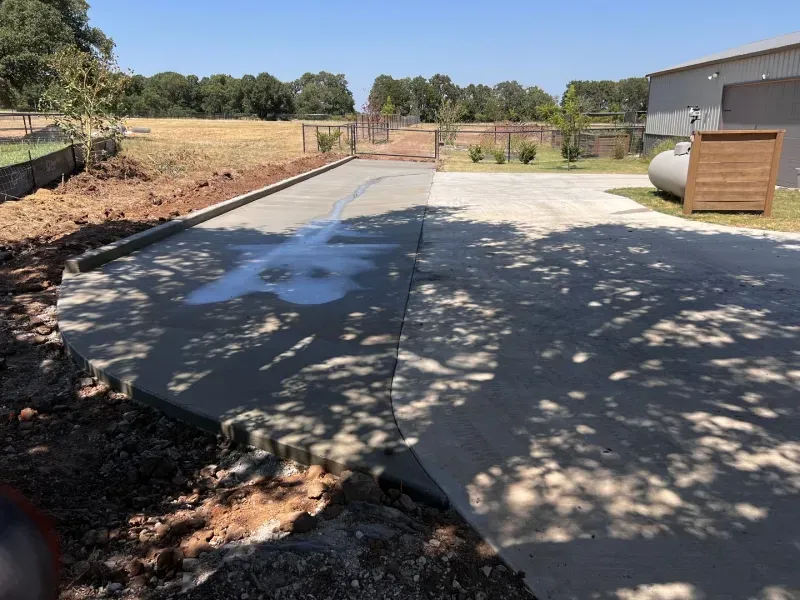 A wet, freshly poured concrete driveway extension next to an older concrete pad on a rural property.