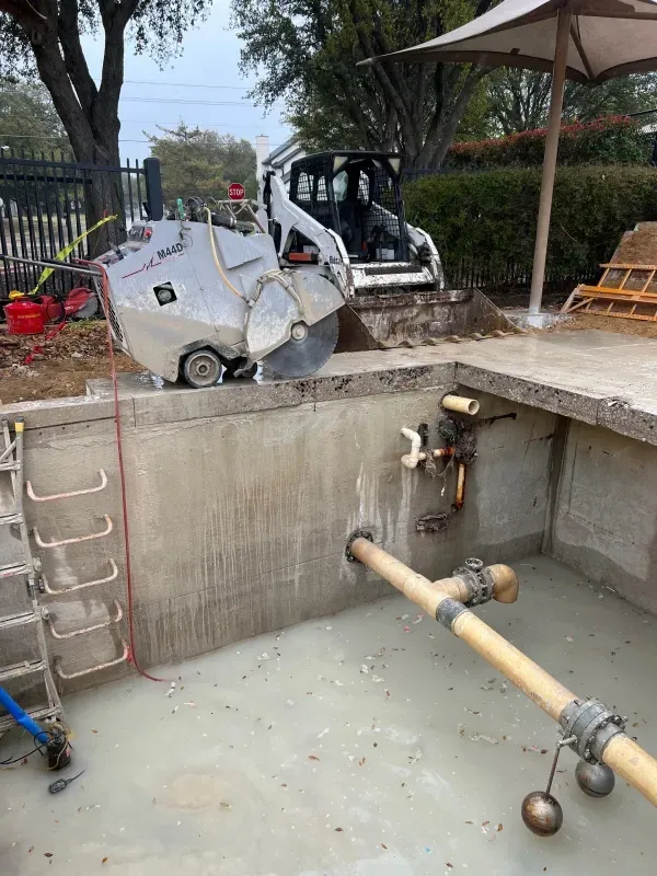 A construction site showing a concrete saw cutting the edge of an empty, unfinished swimming pool.