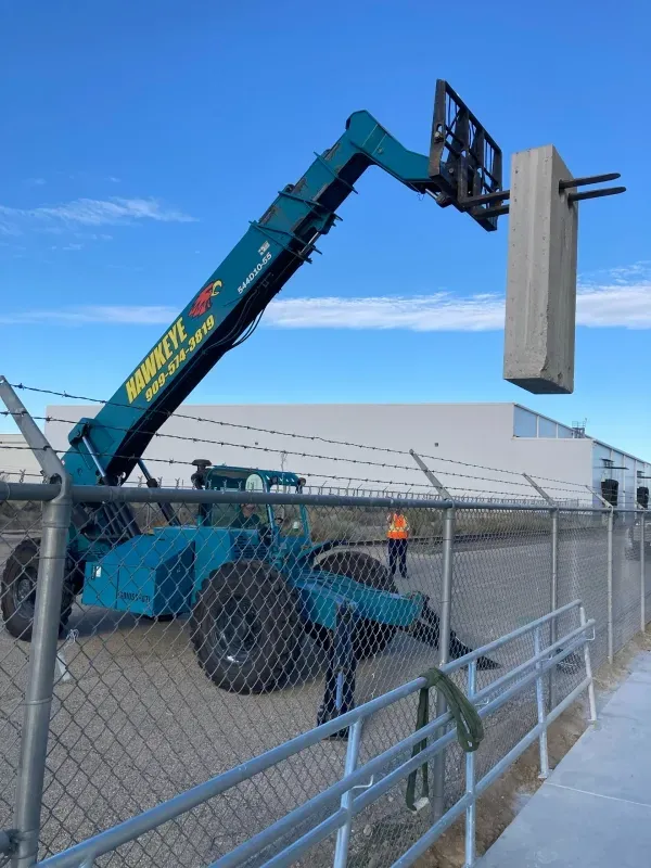 A teal telehandler holds a vertical concrete block outdoors behind a chain-link fence under a clear blue sky.