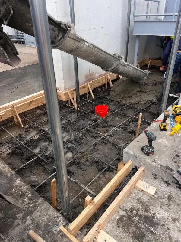 Concrete flows from a chute into a wooden-framed construction area reinforced with a metal wire grid on dirt.
