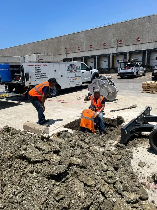 Two workers in high-visibility vests excavate a rectangular pit in a paved lot near a utility truck and a loading dock.