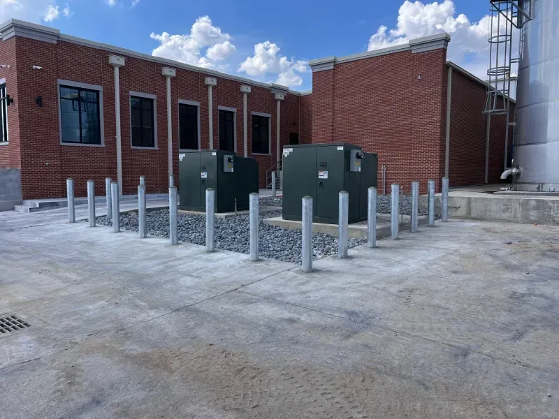 Two dark green electrical transformers sit on a gravel pad, surrounded by a row of protective metal bollards outside.