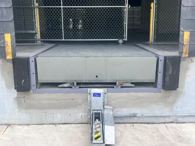 A metal dock leveler centered at a loading bay, flanked by black rubber bumpers on a concrete wall.