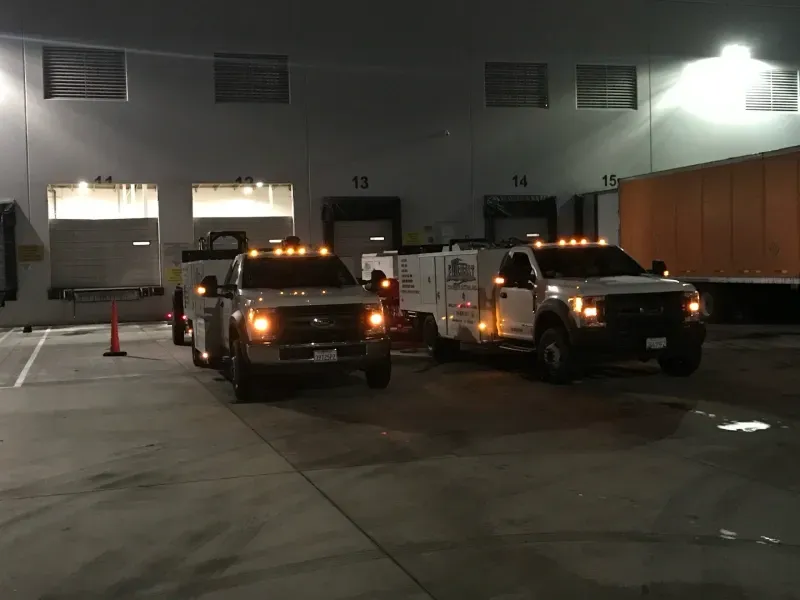 Two white service trucks parked outside loading docks at night under bright overhead lights.