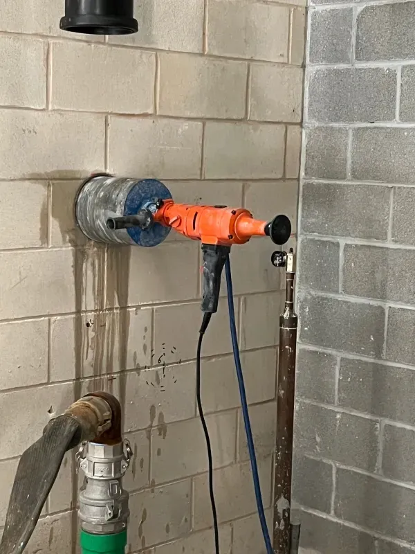 A bright orange core drill is mounted into a tiled wall, with a pipe and water drainage visible below it.