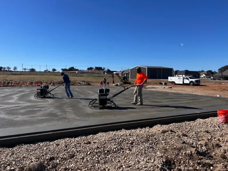 Two workers operate power trowels on a large, freshly poured concrete slab on a sunny, rural construction site.