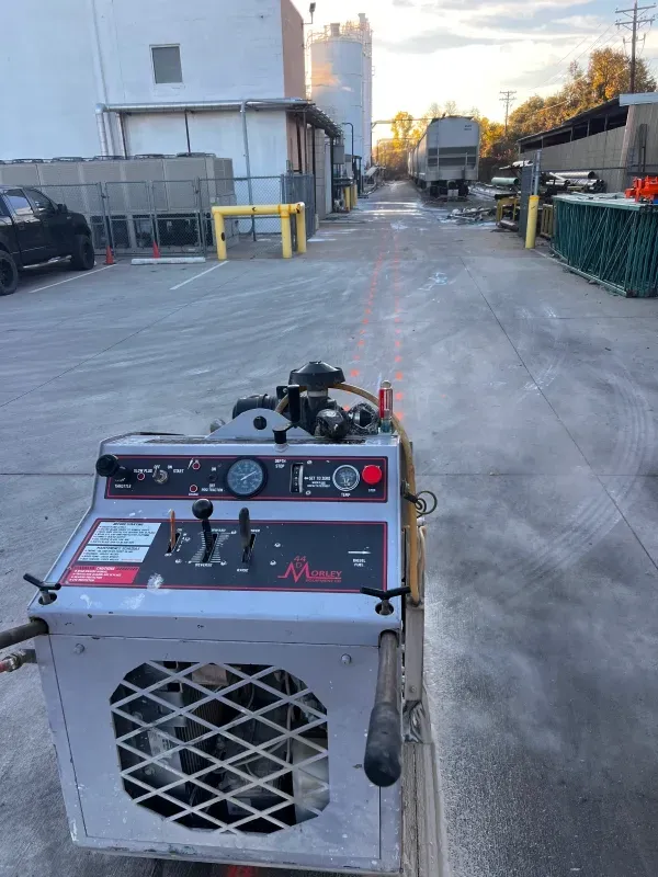 A grey walk-behind floor scrubber in an outdoor parking area, with a white building and a train car in the background.