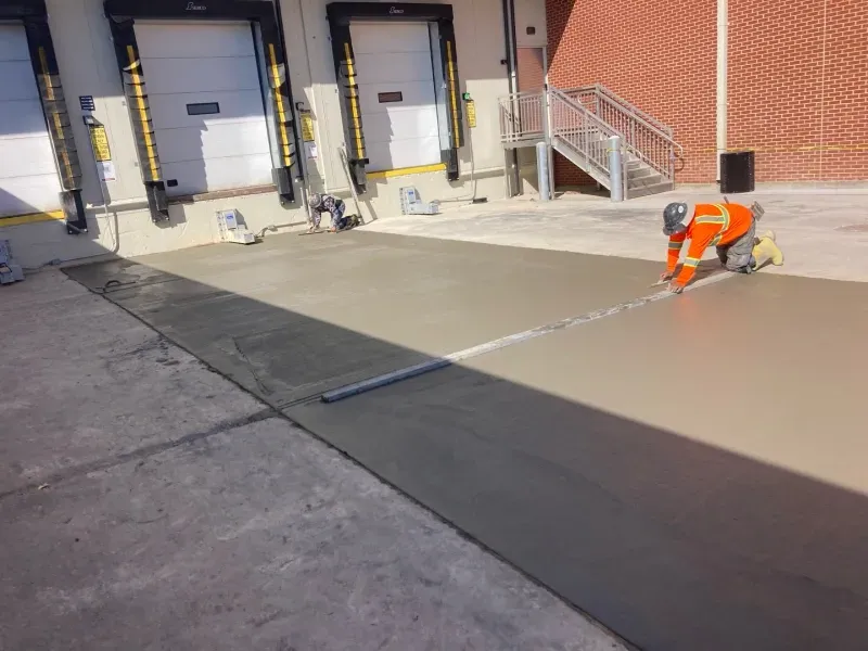 A construction worker in a high-visibility orange jacket smooths wet concrete near a building loading dock.