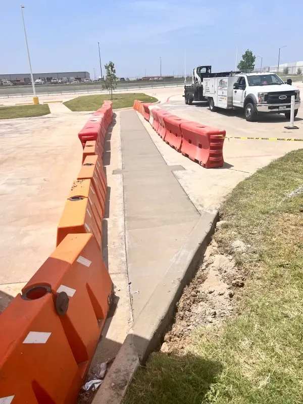 A newly paved concrete path is framed by orange traffic barriers in a parking lot, with a white work truck parked nearby.