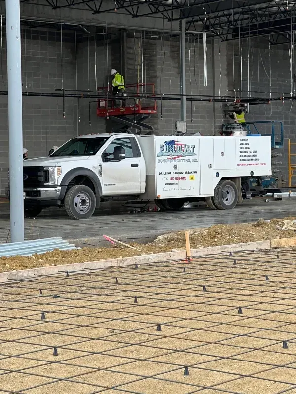 A white service truck parked inside a construction site with workers on lifts and a rebar grid in the foreground.