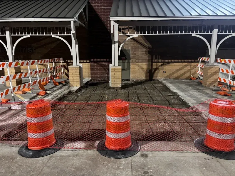 Three orange traffic barrels stand in front of a sidewalk construction zone closed off by red plastic mesh fencing.
