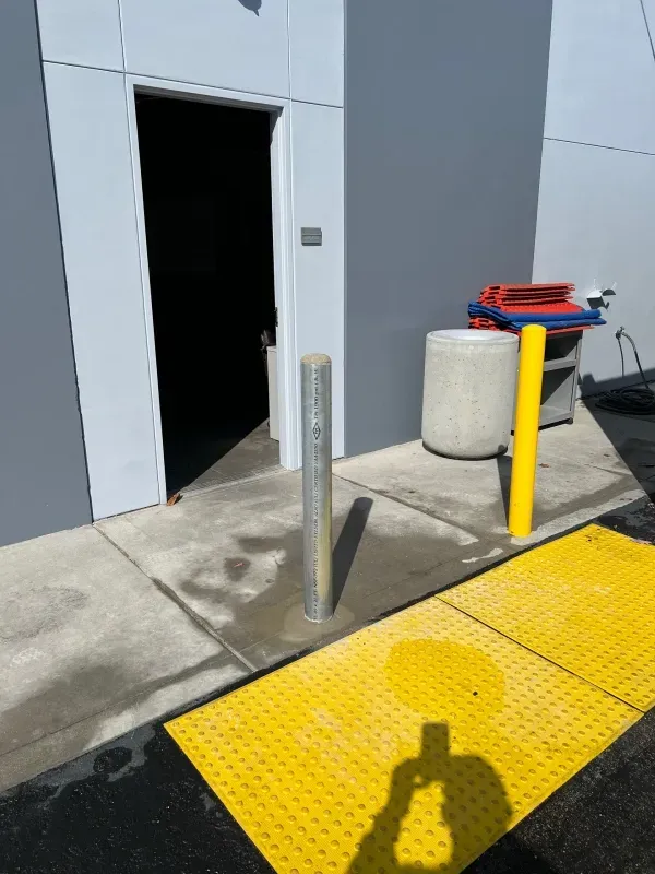 Exterior view of a building entrance with two safety bollards and a yellow tactile paving mat for accessibility.