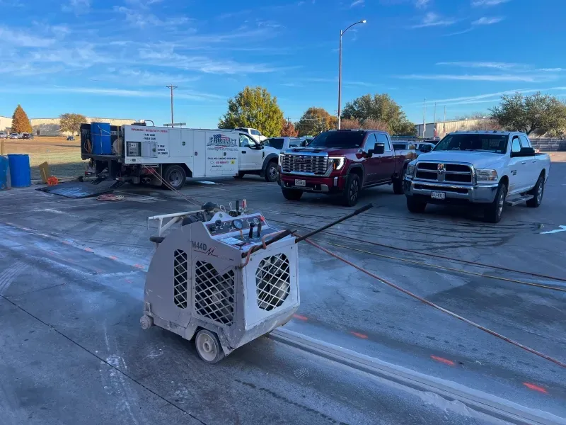 A concrete saw sits on a parking lot next to a utility truck and two pickup trucks under a bright blue sky.