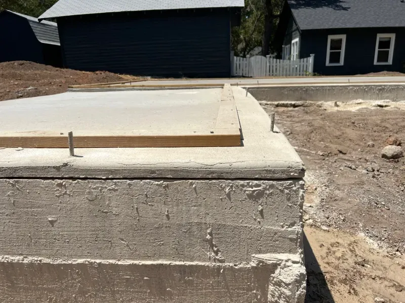 A fresh concrete foundation for a building with metal anchor bolts sticking out, set against a background of houses.