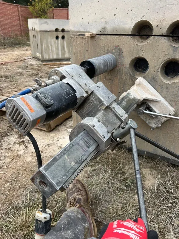 A person uses a core drill mounted on a stand to create circular holes in a large concrete block outdoors.