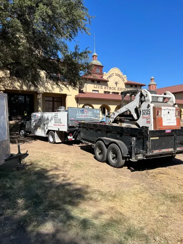 A white service truck pulling a black flatbed trailer with a white Bobcat skid-steer parked in front of a historic building.