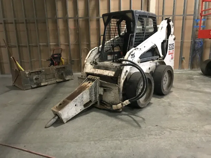 A white Bobcat skid-steer loader equipped with a hydraulic breaker attachment, parked on a concrete floor indoors.