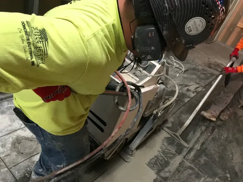 A worker in a bright yellow shirt uses a floor grinder on a concrete surface, with a second person visible in the background.
