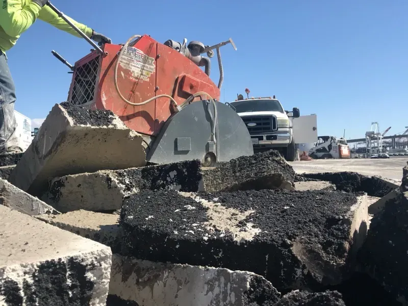 A worker in a high-visibility jacket operates an orange concrete saw on a construction site with broken pavement.