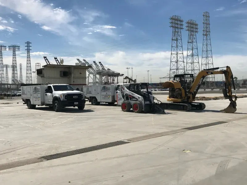 Two service trucks, a skid steer, and an excavator parked on a large concrete lot under a bright, clear sky.