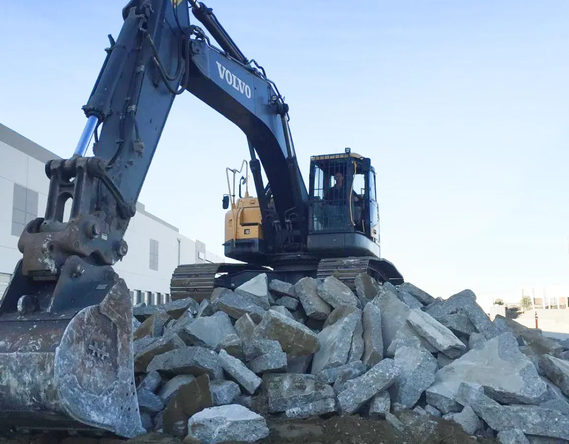 A dark grey Volvo excavator parked on top of a large pile of broken concrete rubble against a clear sky.