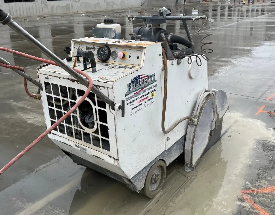 A white walk-behind concrete saw sits on a wet, paved construction site, cutting a groove in the surface.