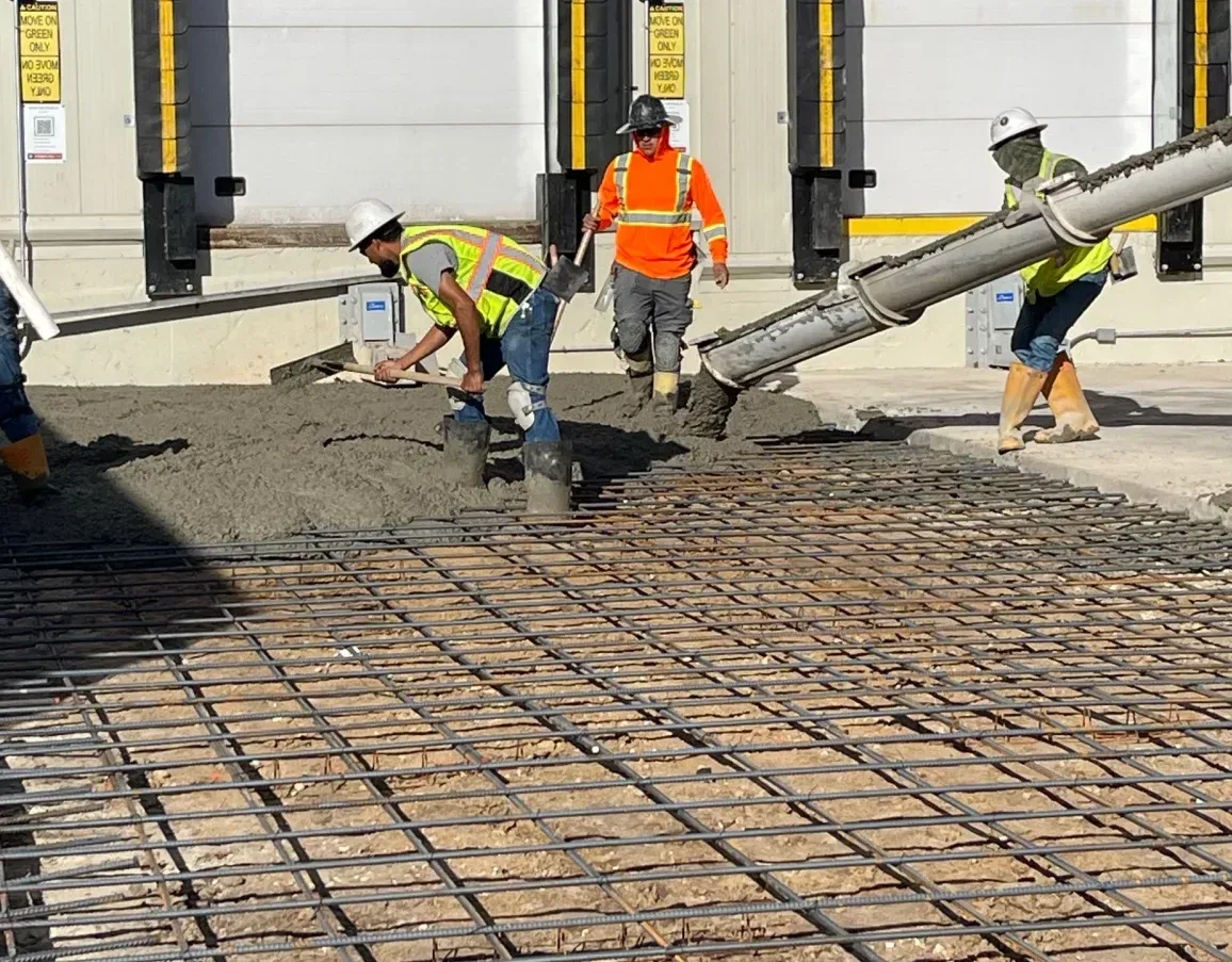 Three construction workers in hard hats and high-visibility vests pour concrete over a rebar grid at a building site.