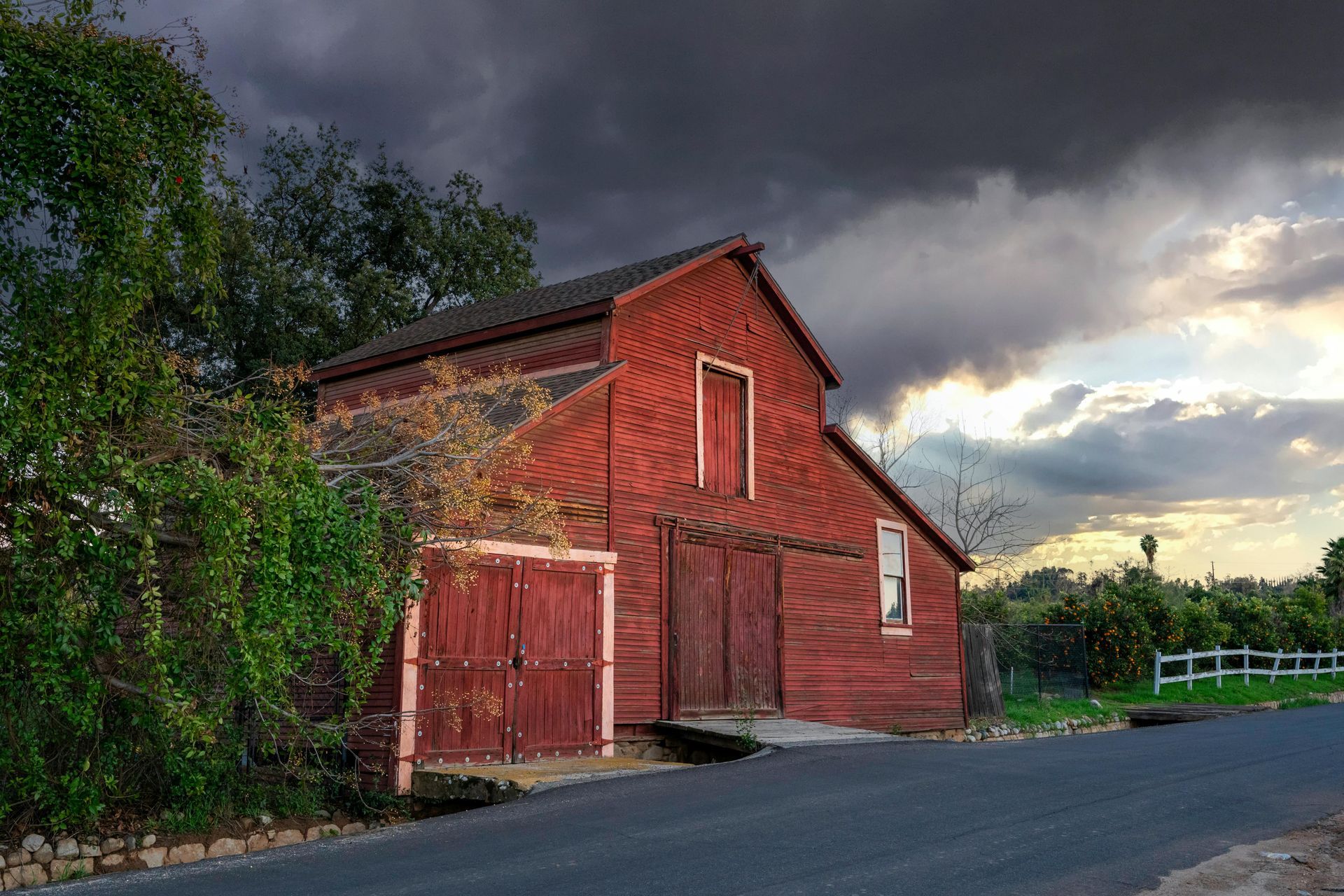 Red barn under a dramatic, stormy sky. Road in foreground, fence to the right.