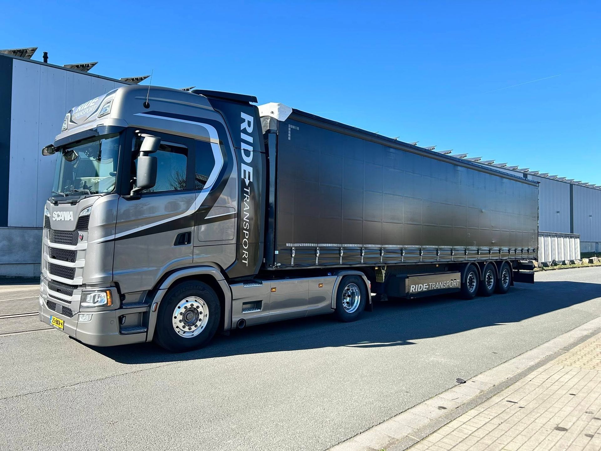 Een grijze vrachtwagen met een zwarte oplegger staat geparkeerd bij een gebouw op een zonnige dag.