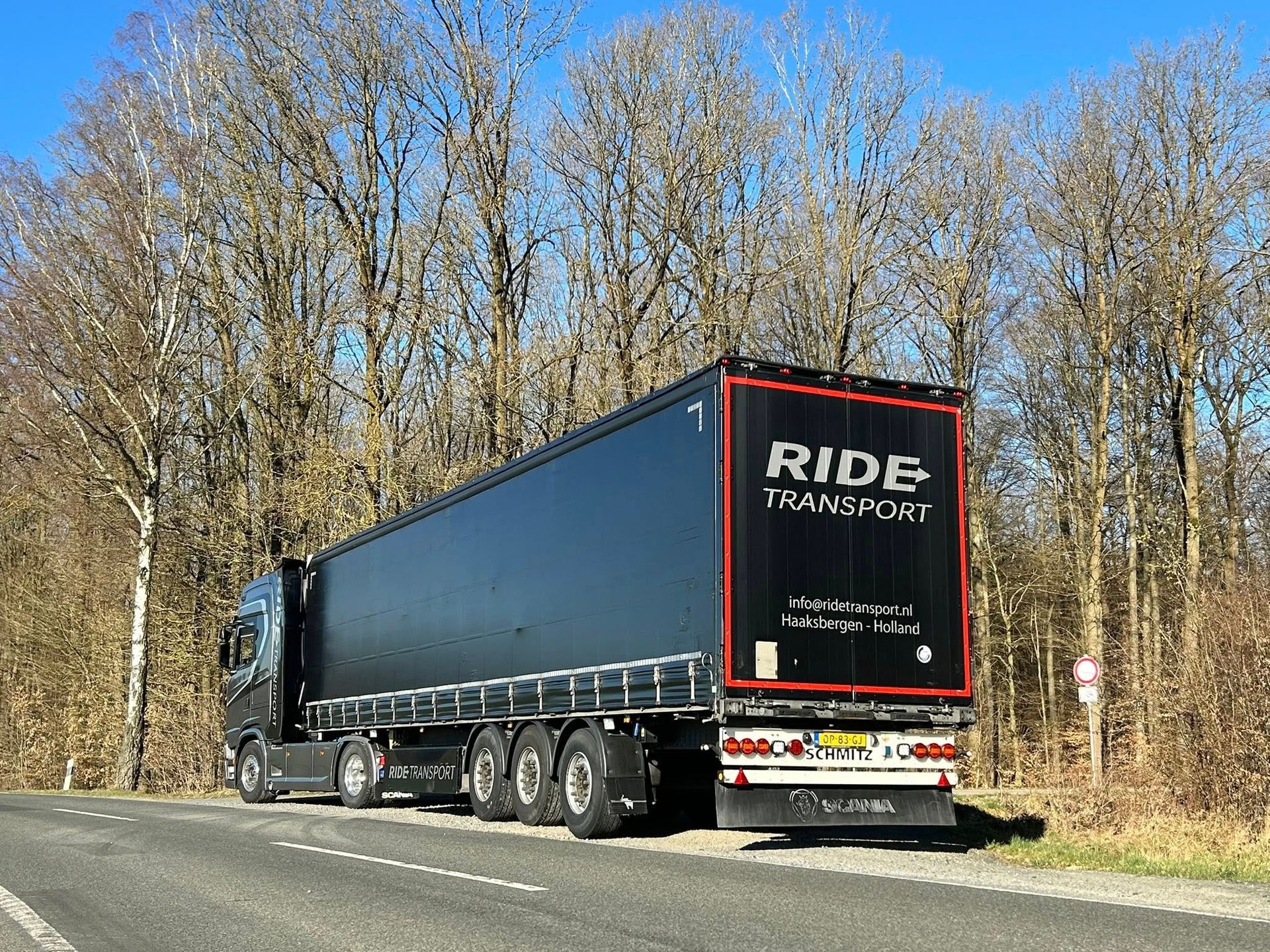 Een zwarte vrachtwagen met oplegger staat geparkeerd op een weg met bomen op de achtergrond onder een blauwe hemel.