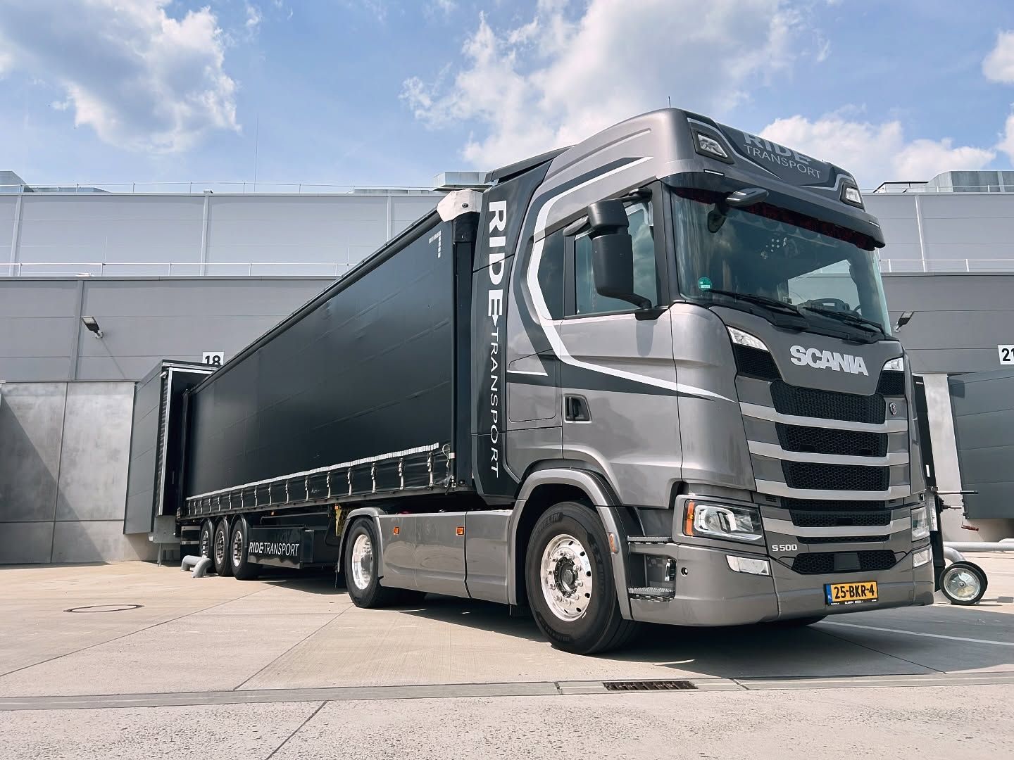 Grijze Scania-vrachtwagen met zwarte oplegger geparkeerd bij een laadperron tegen de achtergrond van de lucht.