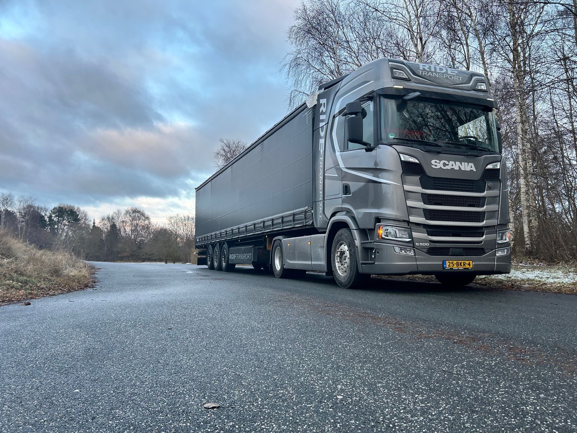 Grijze Scania-vrachtwagen met oplegger op een natte weg, bomen op de achtergrond onder een bewolkte hemel.