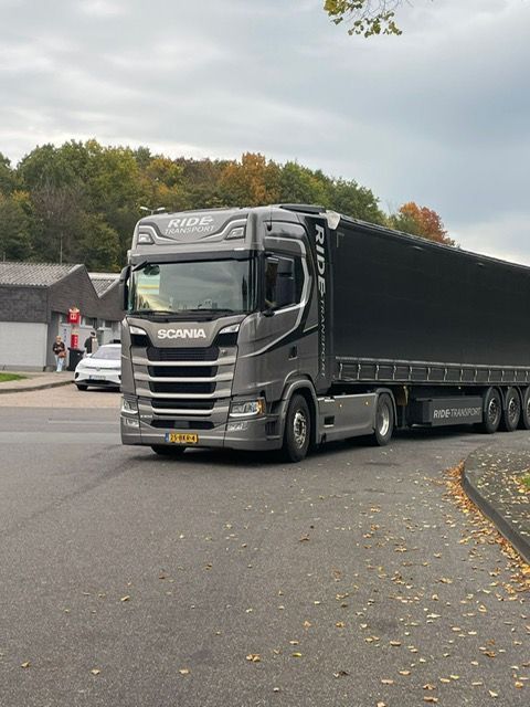 Grijze Scania-vrachtwagen met zwarte oplegger op een weg, met bomen en gebouwen op de achtergrond.