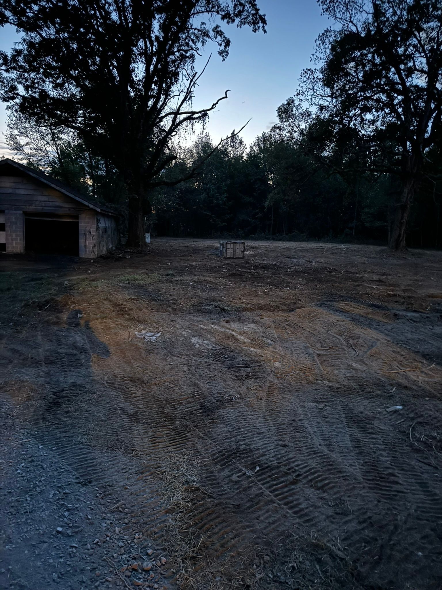 Dirt lot with tractor tracks, shed to the left, trees in background, dusk.