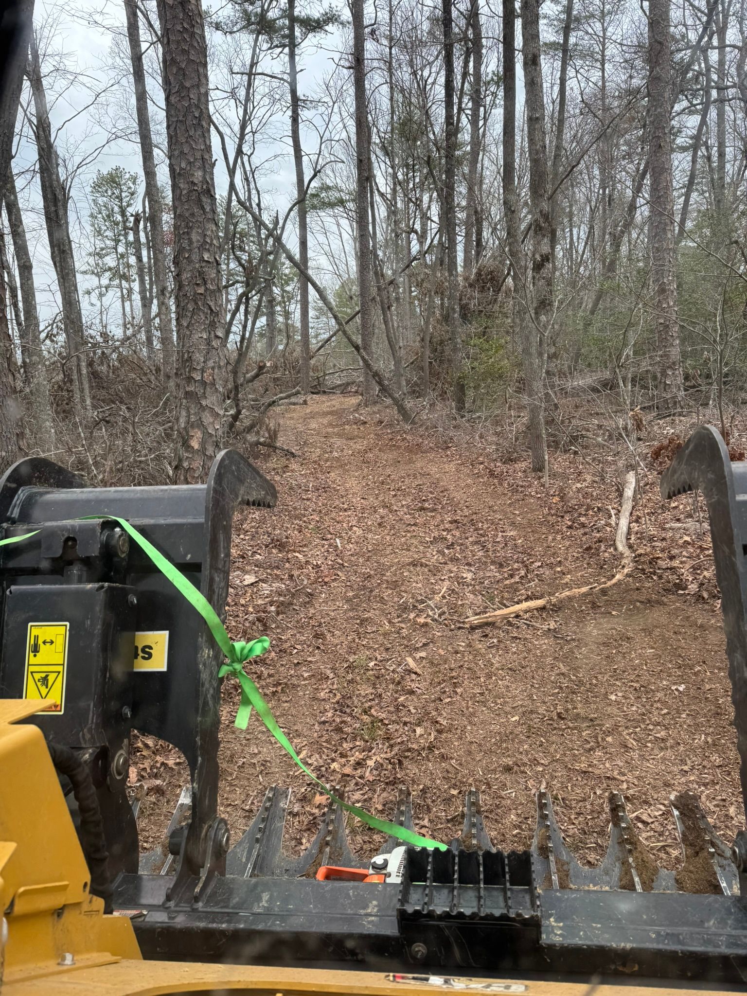 View from a vehicle's bucket: A leaf-covered path winds through a forest. A green strap is attached to the bucket.